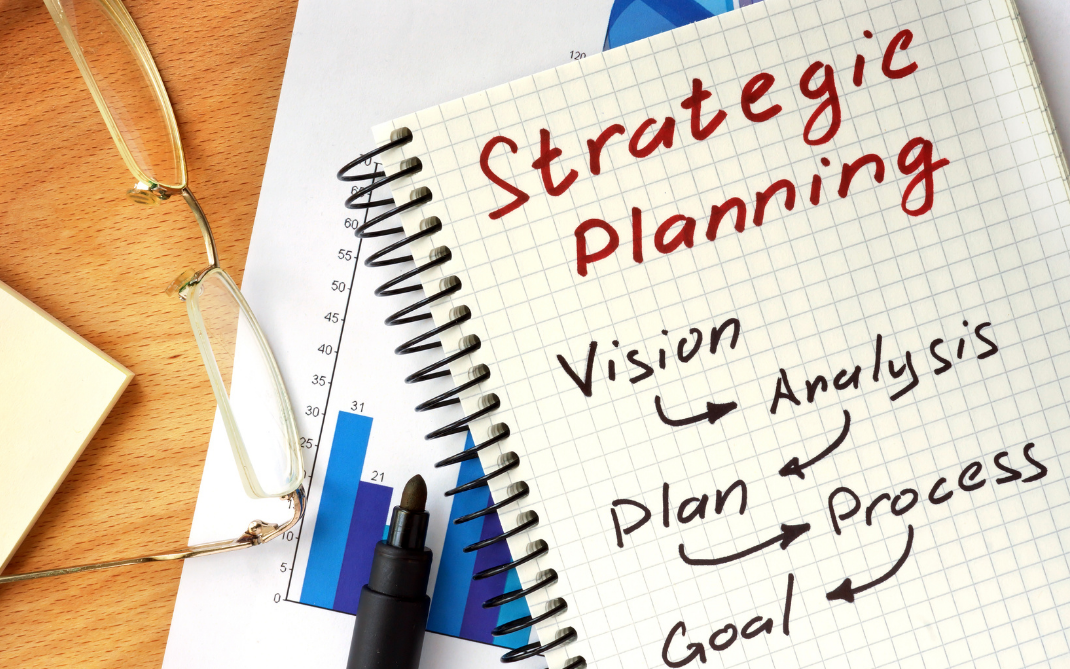 background image of notebook on desk with pens and other paper. written on notepad are the words Strategic Planning