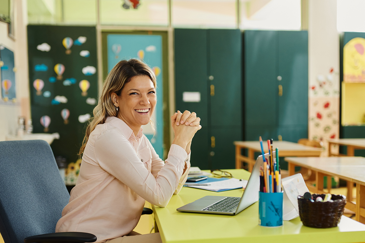 smiling teacher sitting at desk with computer enjoying job in education