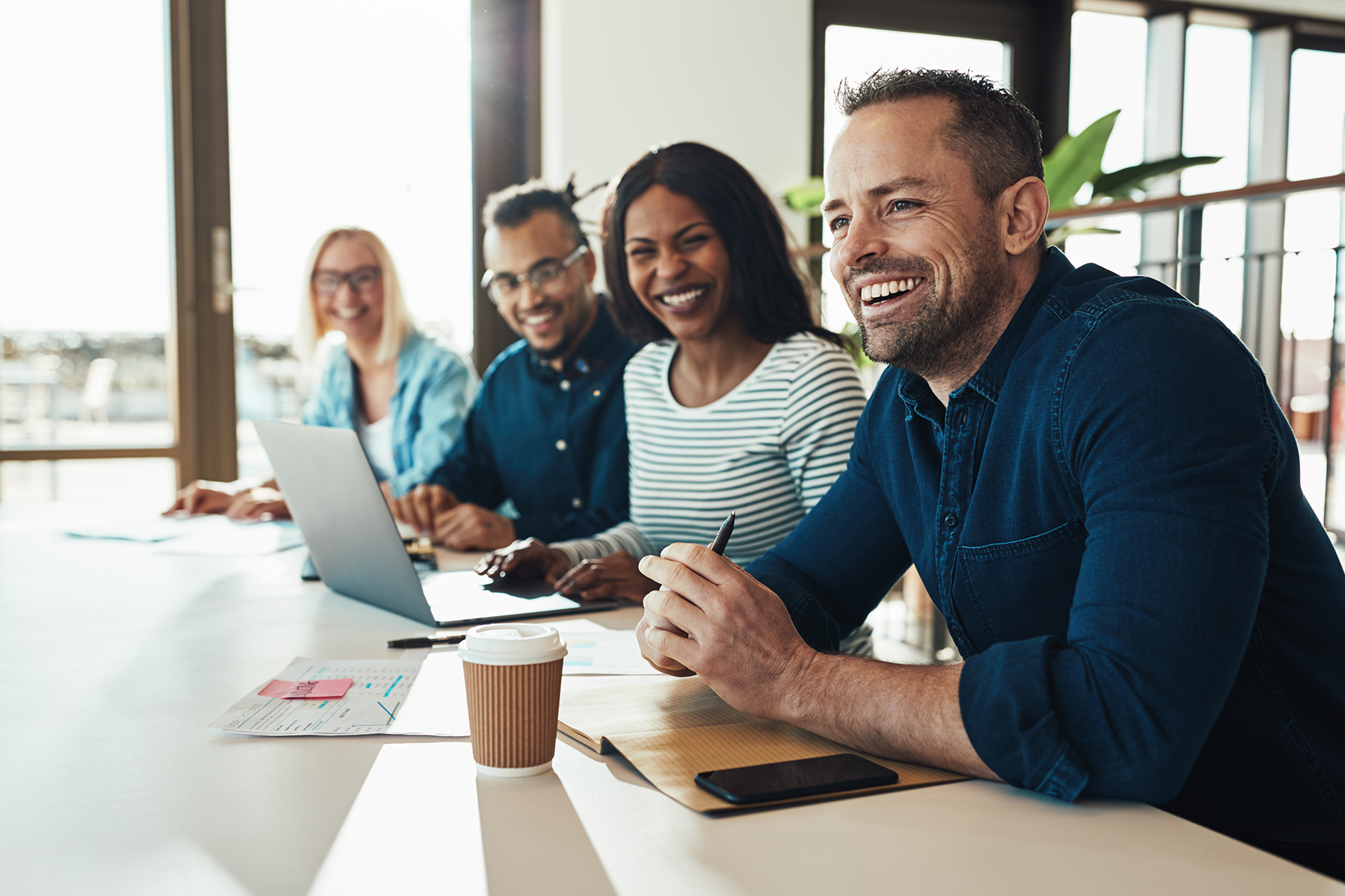 Diverse group of young businesspeople laughing while sitting together in a row at an office desk during a meeting