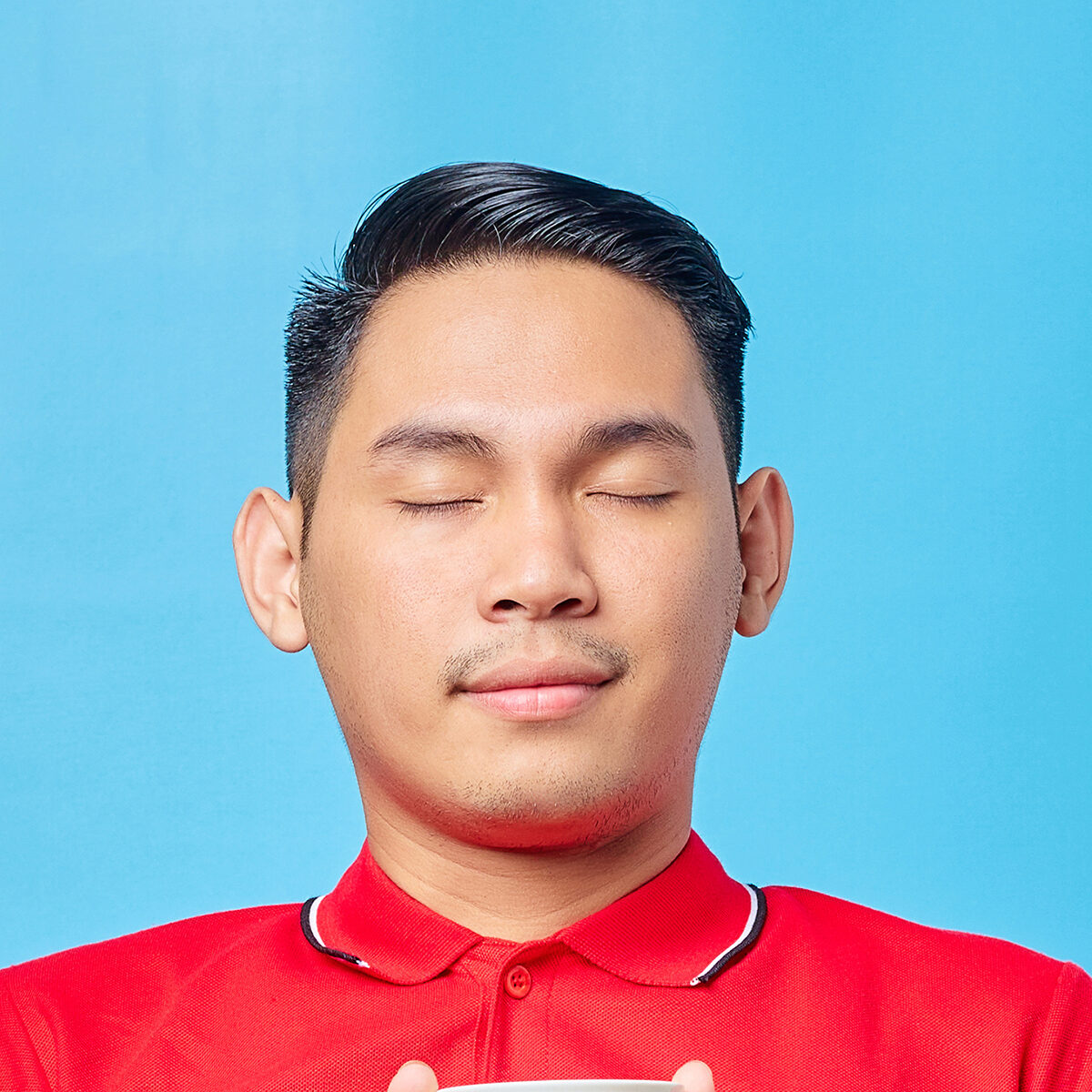 Portrait of handsome Asian young man holding cup of coffee drink and closing his eyes while inhaling delicious coffee aroma isolated on blue background