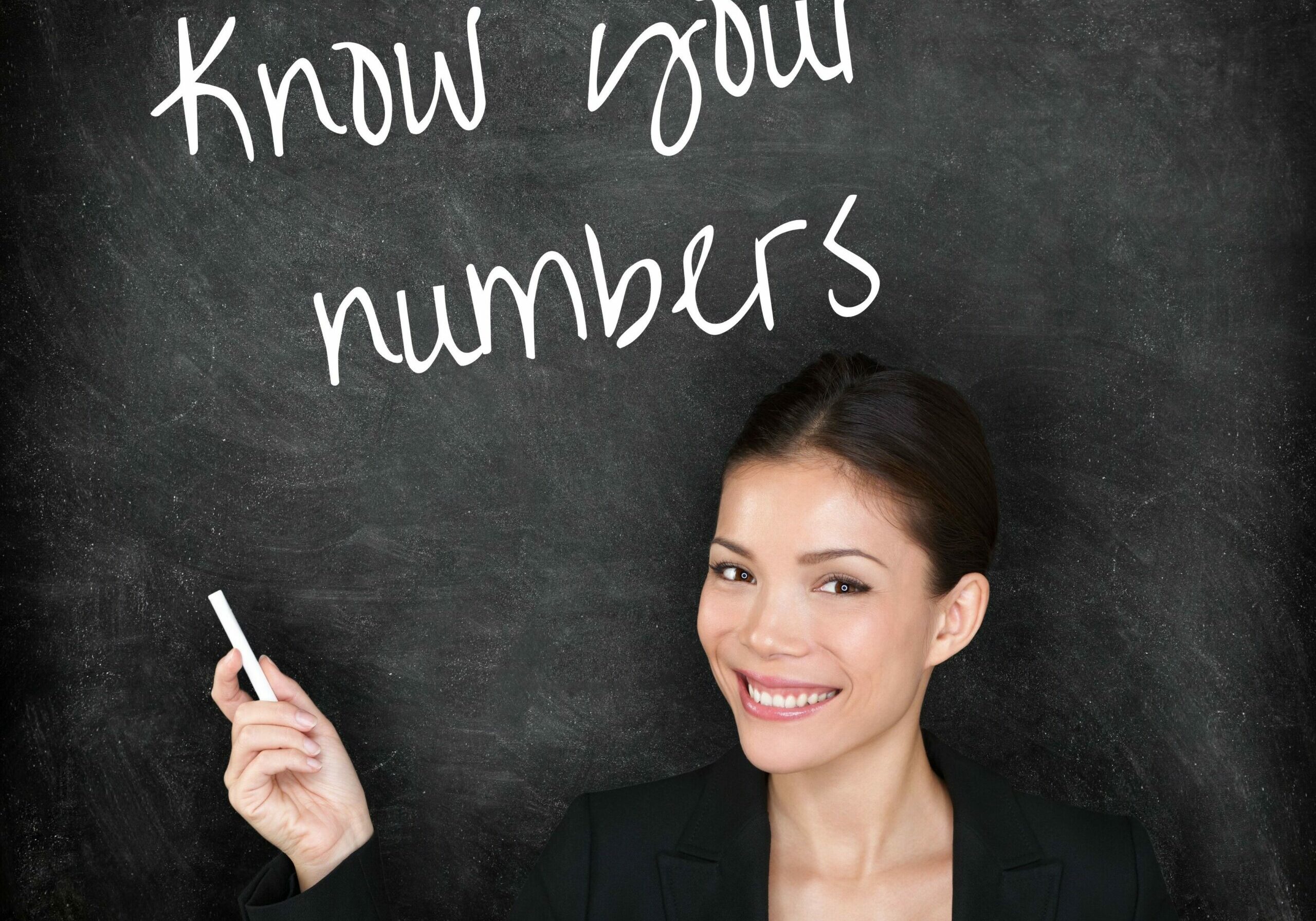 smiling young woman facing camera with back to chalkboard and chalk in hand. She has written 'know your numbers' on chalkboard.